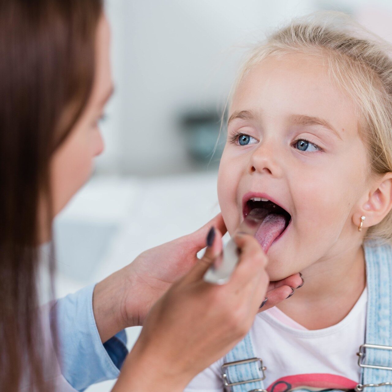 cute-little-blond-girl-opening-mouth-while-sitting-front-clinician-examining-her-throat-with-stell-spatula-min